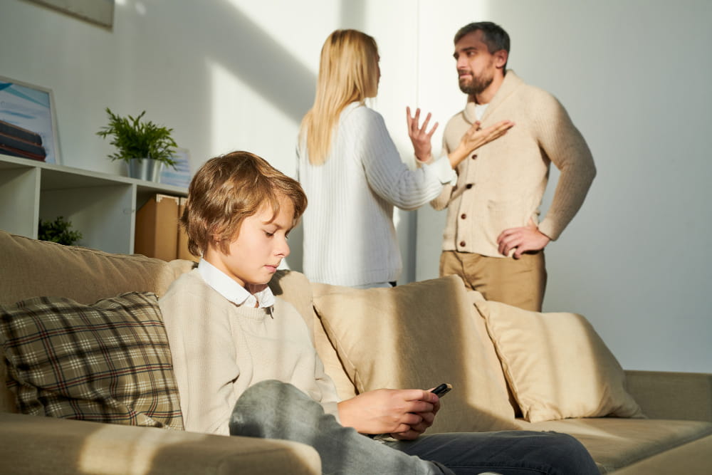 Enfant assis sur un canapé regardant son téléphone pendant que ses parents se disputent en arrière-plan, illustrant les tensions liées à la garde des enfants.